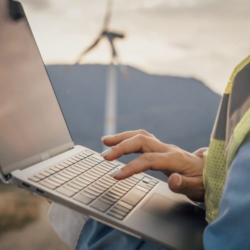 A professional in a safety vest uses a laptop outdoors among wind turbines, showcasing the blend of modern green tech and remote work within natural settings, balancing innovation and nature