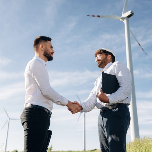 Handshake, successful deal. Two men engineers are in field, with wind turbines.