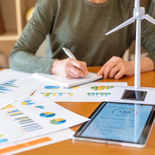 Woman focusing on data analysis and planning for green business and renewable energy, reviewing performance charts on papers and a tablet with a wind turbine model on a wooden office desk